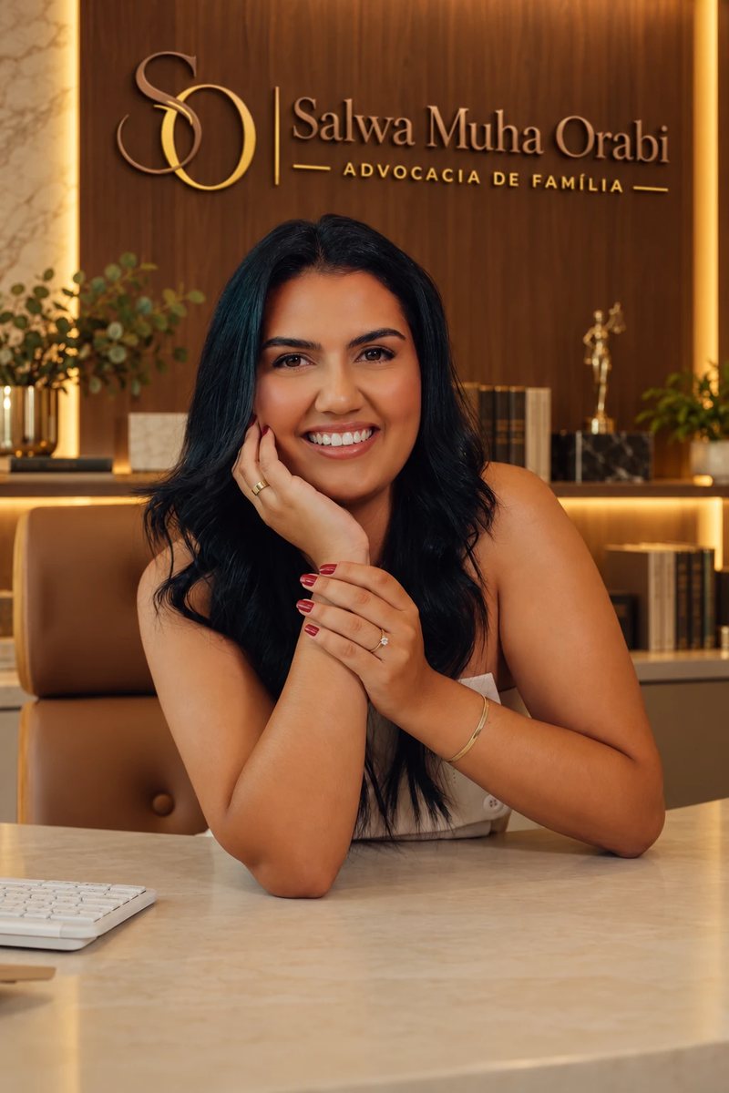 Retrato de Salwa Muha Orabi, advogada, sorrindo, com o rosto apoiado na mão, em seu escritório, com o logotipo SO Salwa Muha Orabi Advocacia de Família na parede ao fundo.
